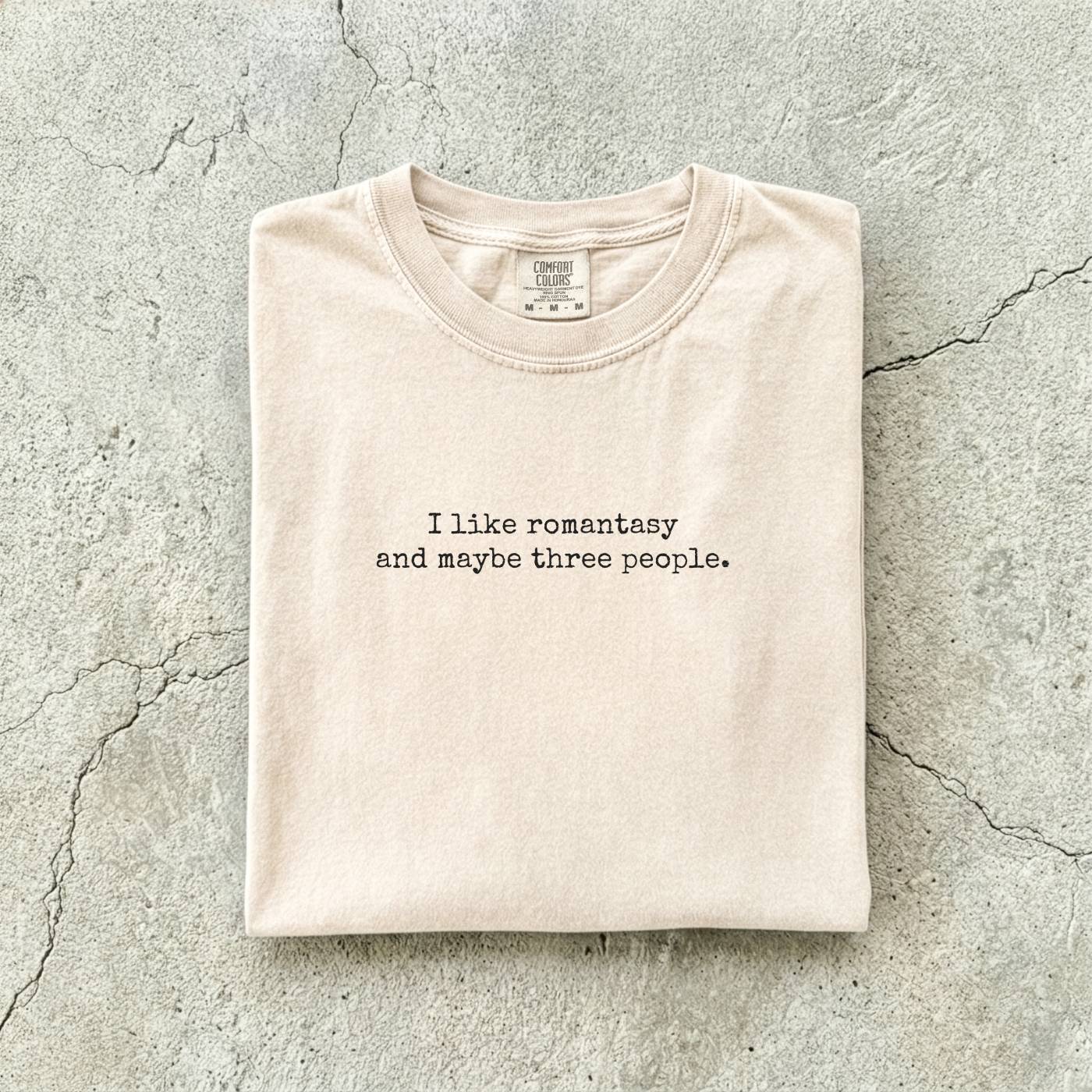 Ivory Comfort Colors tee folded flat reading "I like romantasy and maybe three people." in a typewriter font.