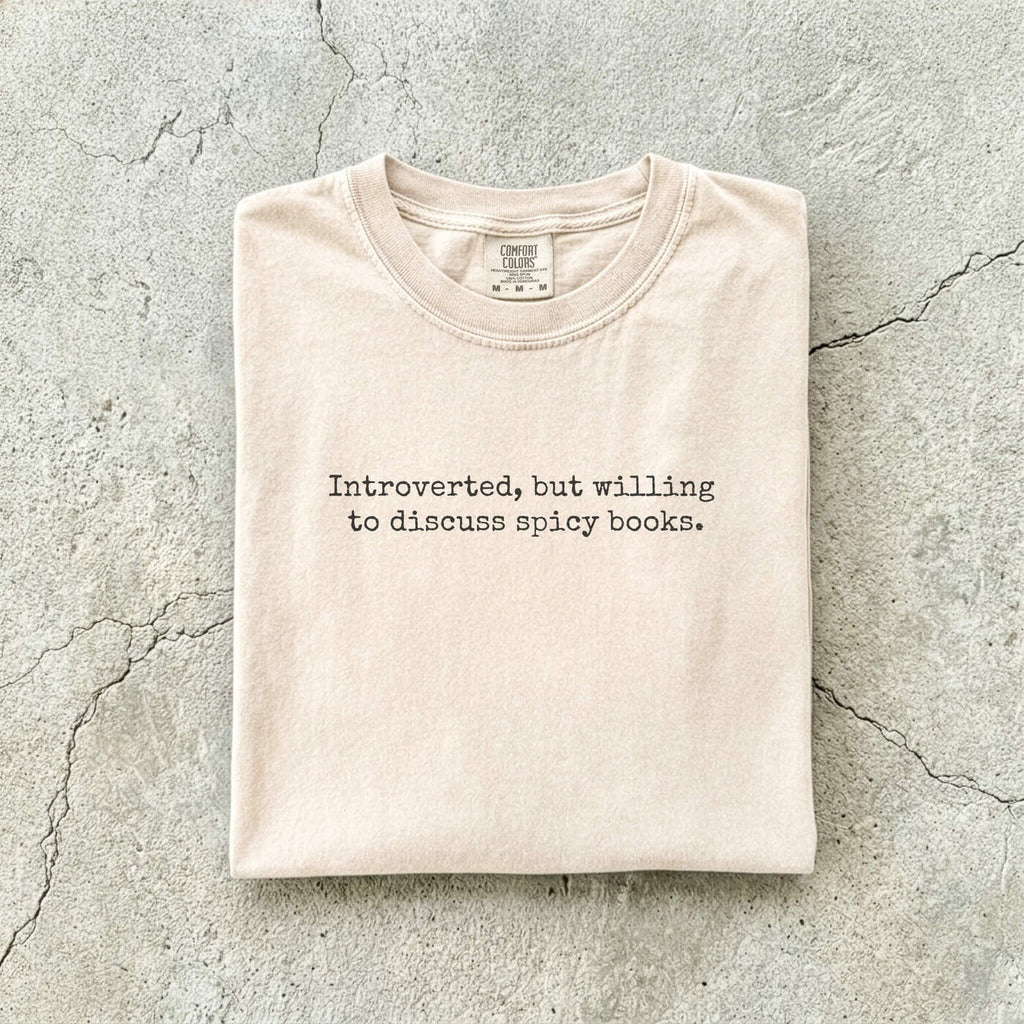 Ivory Comfort Colors tee folded flat on a concrete background reading "Introverted, but willing to discuss spicy books." in a minimal typewriter font.