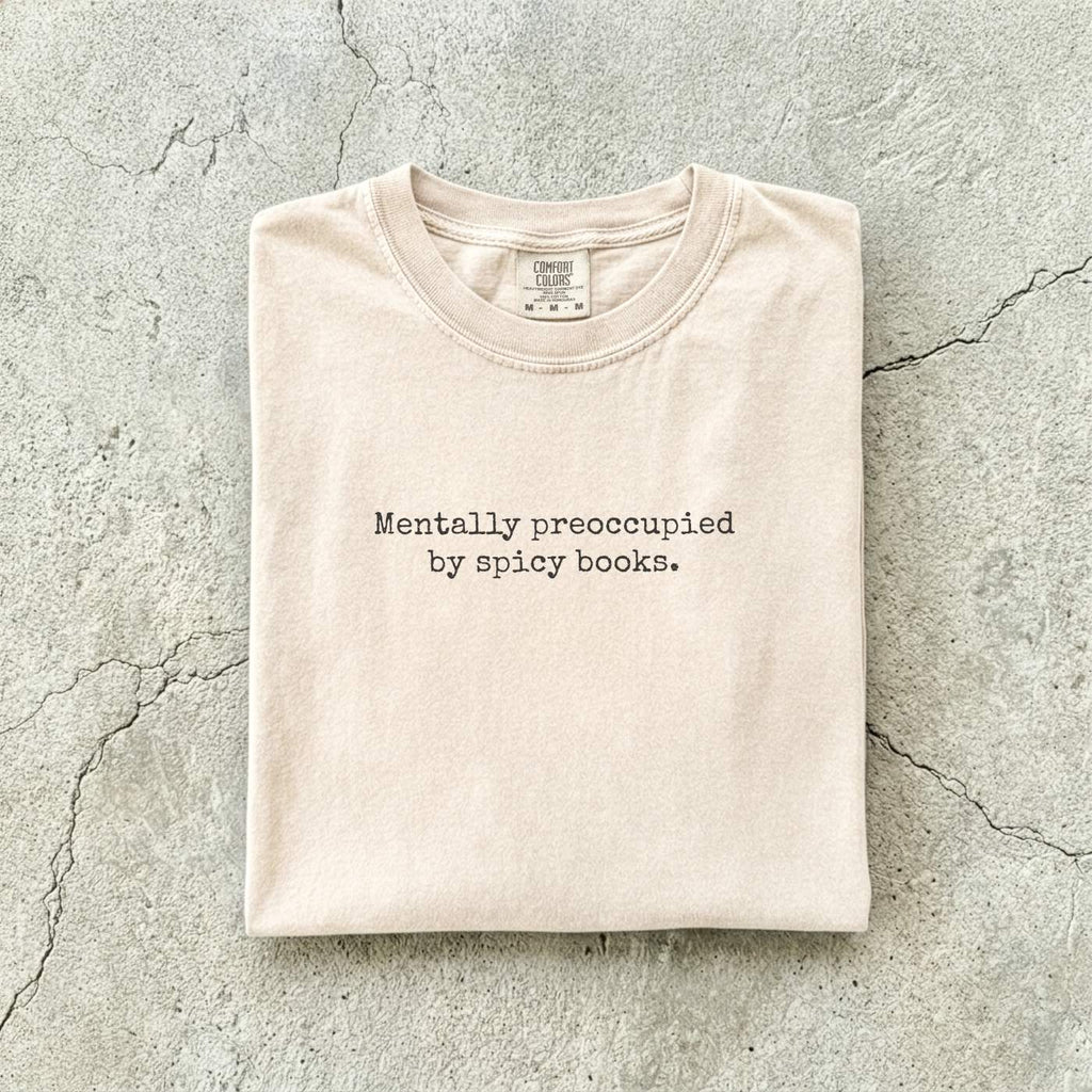 Ivory Comfort Colors tee folded flat on a concrete background reading "Mentally preoccupied by spicy books." in a minimalist typewriter font.