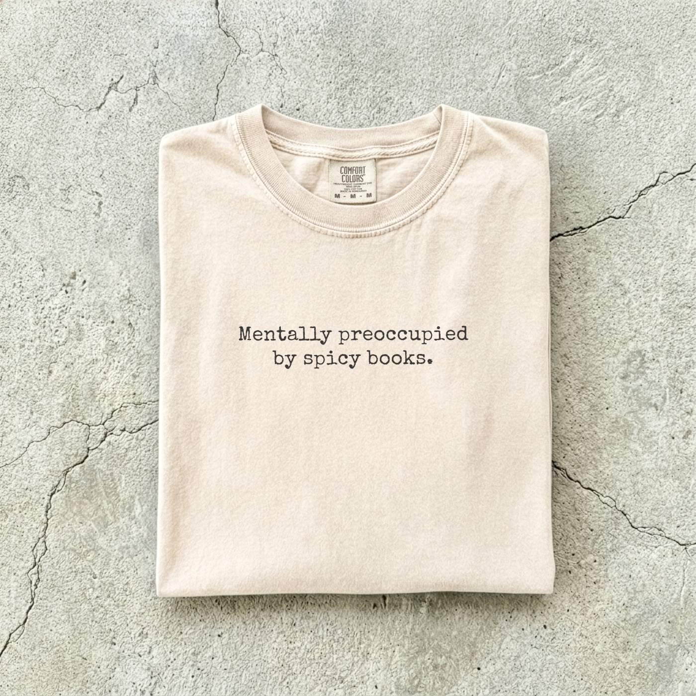 Ivory Comfort Colors tee folded flat on a concrete background reading "Mentally preoccupied by spicy books." in a minimalist typewriter font.