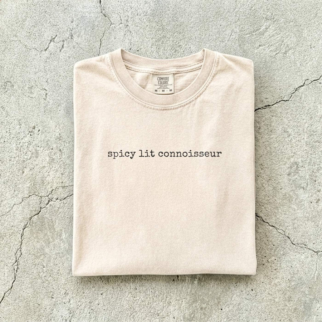 Ivory Comfort Colors tee folded flat on a concrete background reading "spicy lit connoisseur" in a minimal typewriter font.