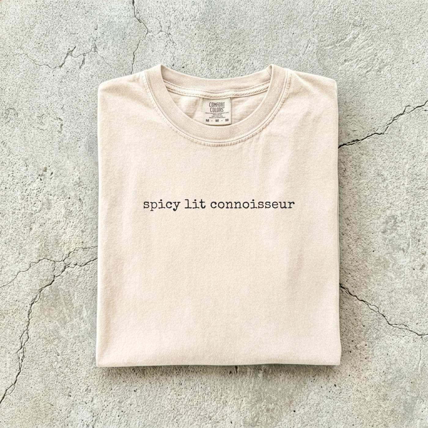 Ivory Comfort Colors tee folded flat on a concrete background reading "spicy lit connoisseur" in a minimal typewriter font.