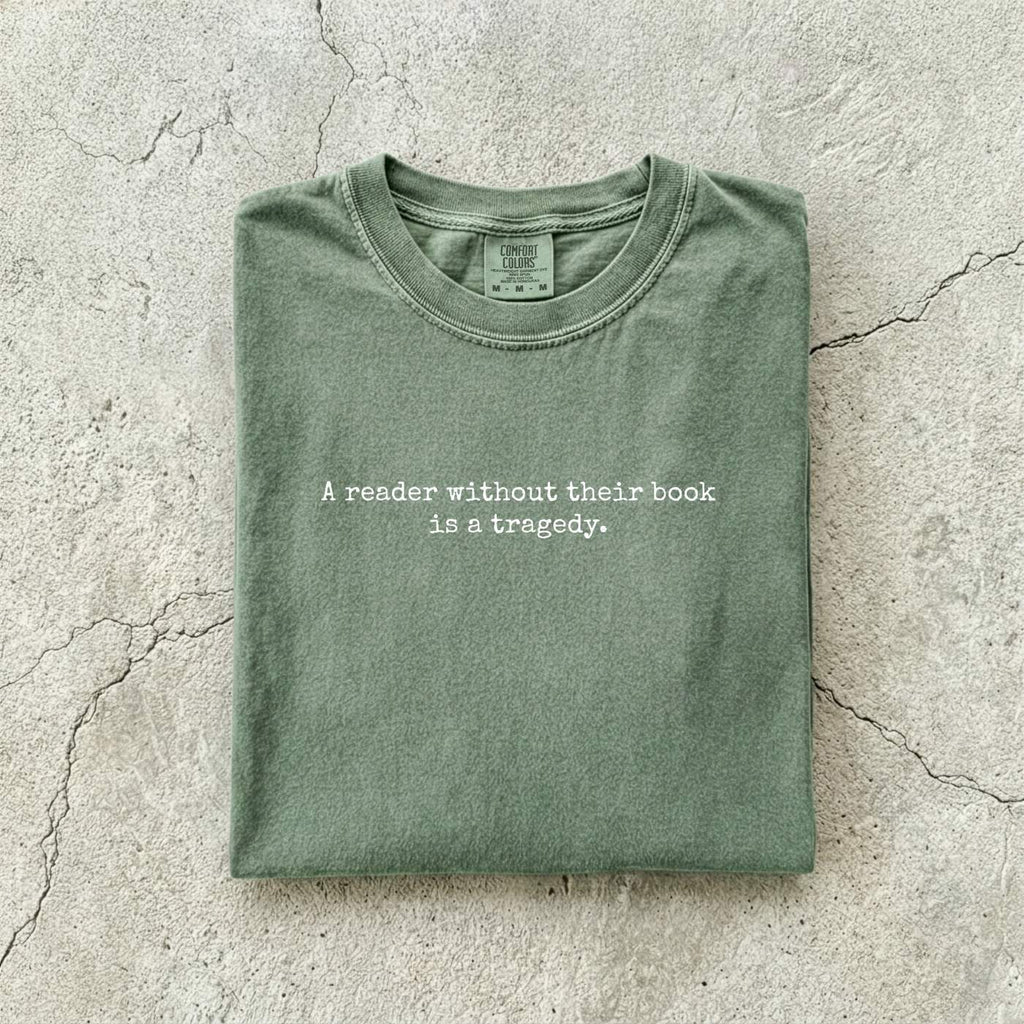 Moss Comfort Colors tee folded flat on a concrete background reading "A reader without their book is a tragedy." in a minimal typewriter font.
