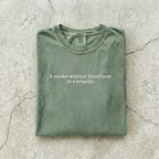 Moss Comfort Colors tee folded flat on a concrete background reading "A reader without their book is a tragedy." in a minimal typewriter font.