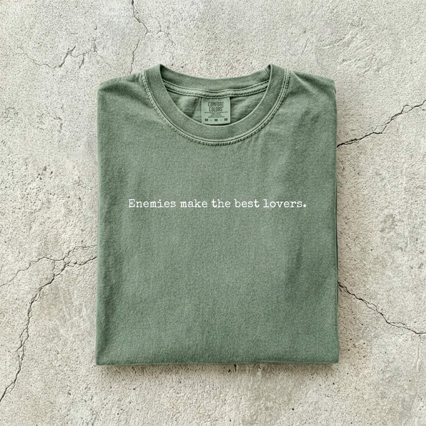 Moss Comfort Colors tee folded flat on a concrete background reading "Enemies make the best lovers." in a minimal typewriter font.