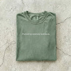 Moss Comfort Colors tee folded flat on a concrete background reading "Fueled by anxiety and smut." in minimal typewriter font.