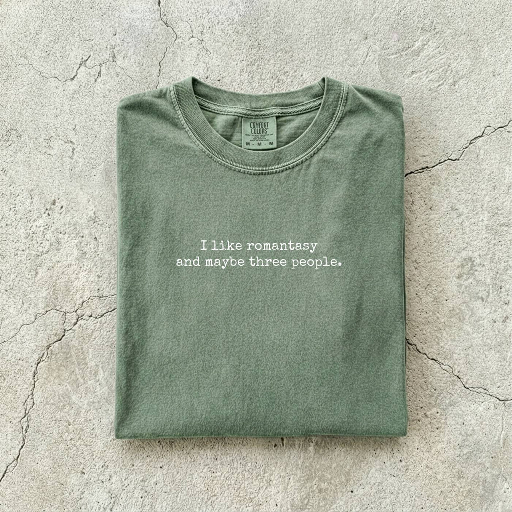 Moss Comfort Colors tee folded flat reading "I like romantasy and maybe three people." in a typewriter font.