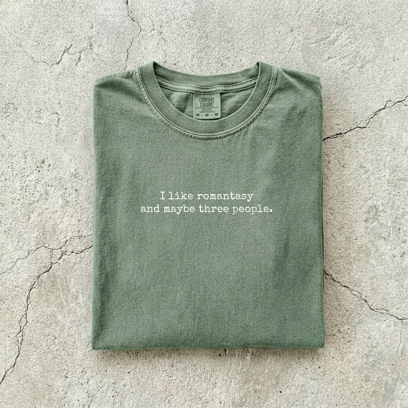 Moss Comfort Colors tee folded flat reading "I like romantasy and maybe three people." in a typewriter font.