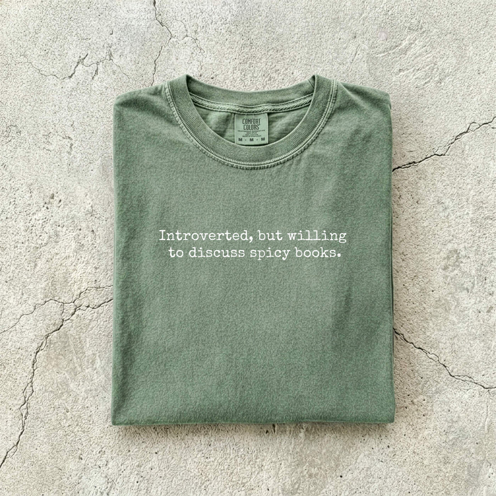 Moss Comfort Colors tee folded flat on a concrete background reading "Introverted, but willing to discuss spicy books." in a minimal typewriter font.