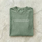 Moss Comfort Colors tee folded flat on a concrete background reading "Introverted, but willing to discuss spicy books." in a minimal typewriter font.