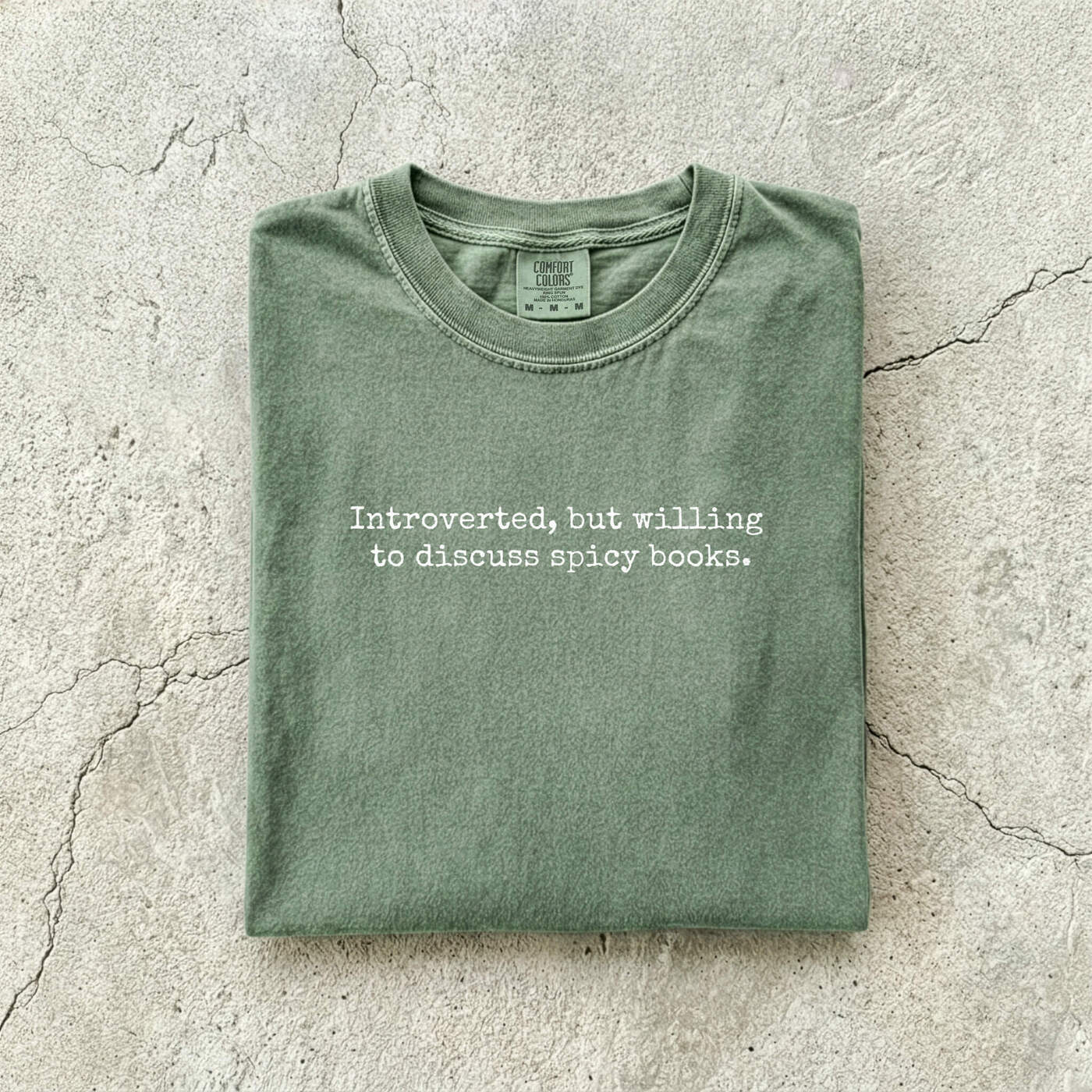 Moss Comfort Colors tee folded flat on a concrete background reading "Introverted, but willing to discuss spicy books." in a minimal typewriter font.