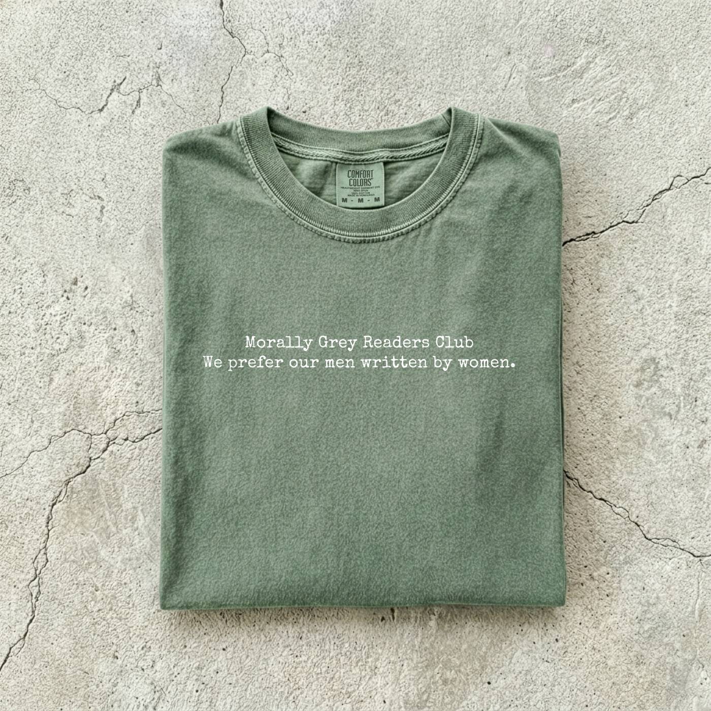 Moss Comfort Colors tee folded flat on a concrete background reading "Morally grey readers club. We prefer our men written by women." in a minimal typewriter font.
