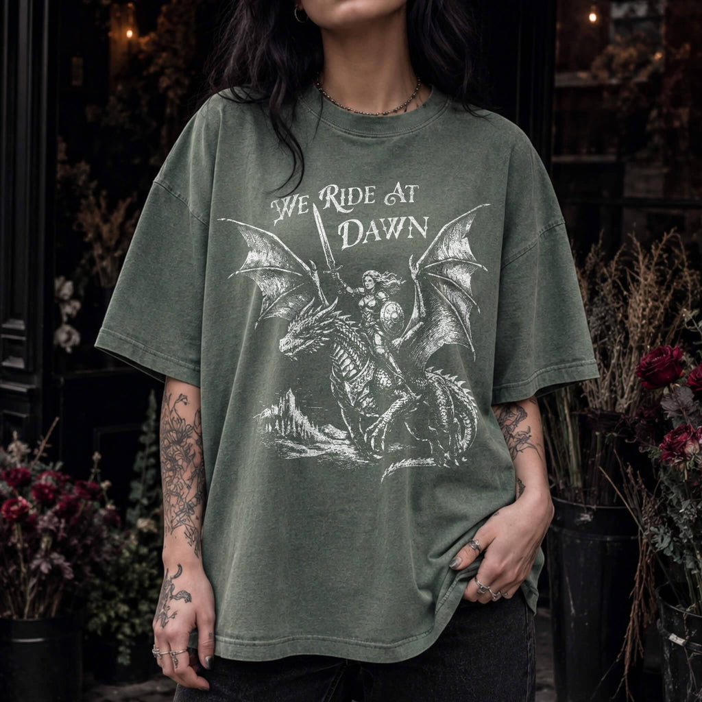Woman wearing a moss color relaxed fit Comfort Colors tee with a vintage graphic of a woman warrior dragon rider and "We Ride at Dawn" text.