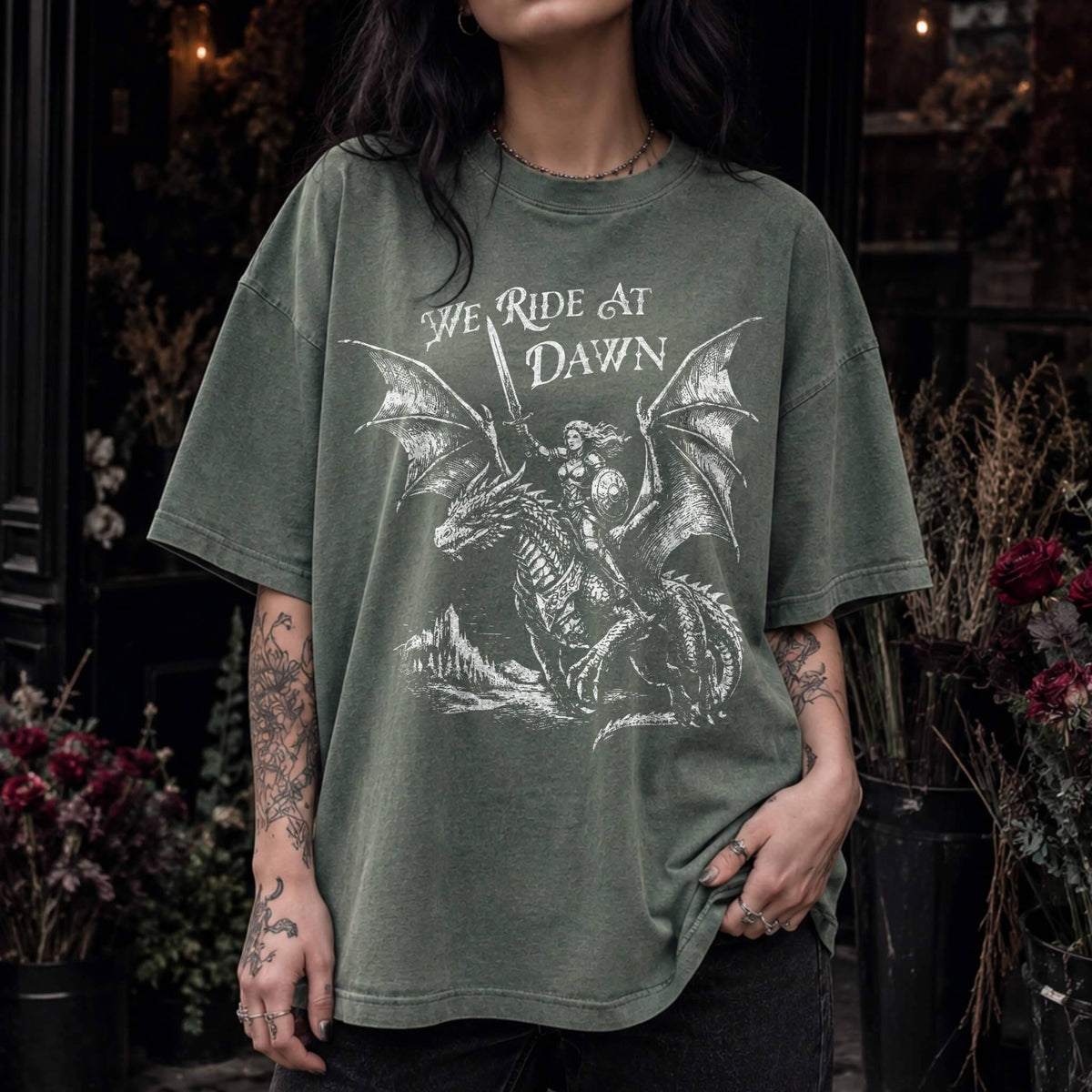 Woman wearing a moss color relaxed fit Comfort Colors tee with a vintage graphic of a woman warrior dragon rider and "We Ride at Dawn" text.