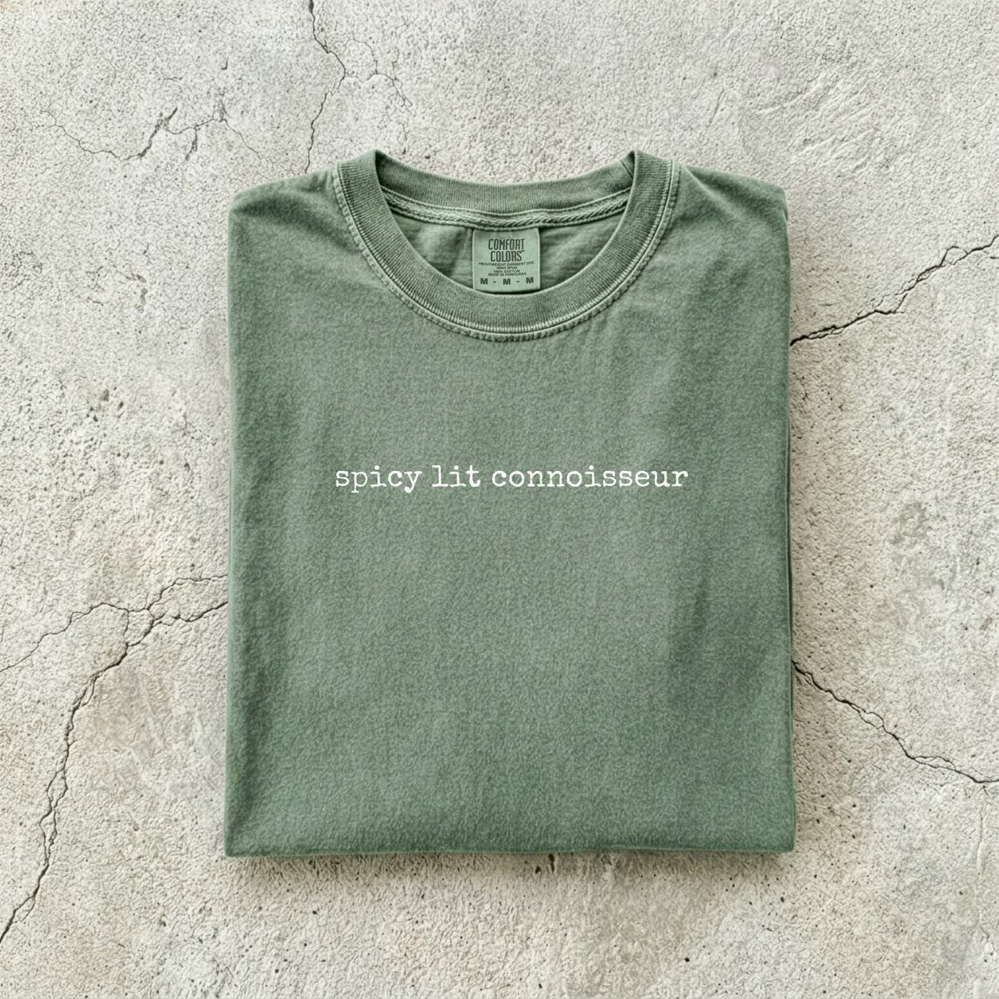 Moss Comfort Colors tee folded flat on a concrete background reading "spicy lit connoisseur" in a minimal typewriter font.