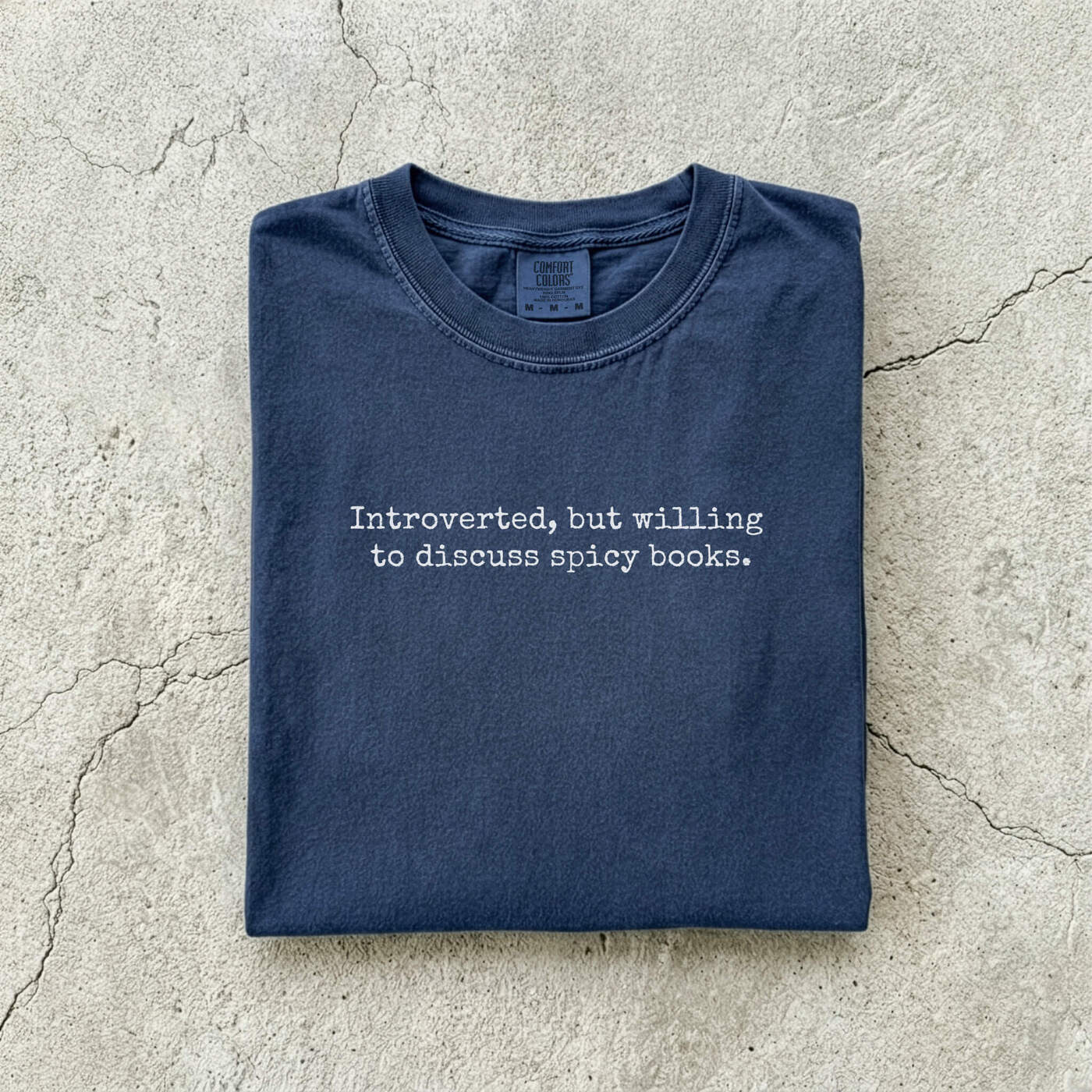 Navy Comfort Colors tee folded flat on a concrete background reading "Introverted, but willing to discuss spicy books." in a minimal typewriter font.