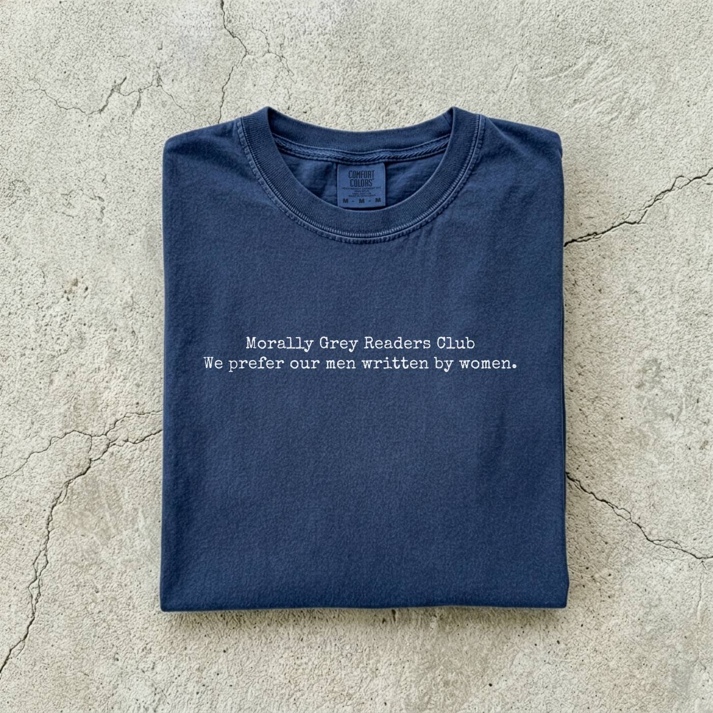 Navy Comfort Colors tee folded flat on a concrete background reading "Morally grey readers club. We prefer our men written by women." in a minimal typewriter font.