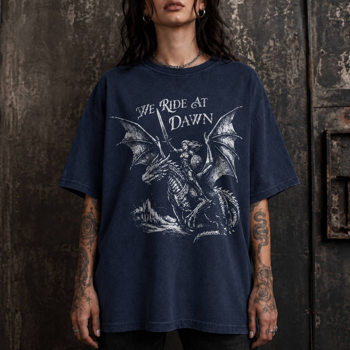 Woman wearing a navy color relaxed fit Comfort Colors tee with a vintage graphic of a woman warrior dragon rider and "We Ride at Dawn" text.