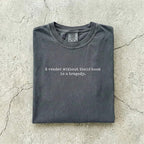 Pepper Comfort Colors tee folded flat on a concrete background reading "A reader without their book is a tragedy." in a minimal typewriter font.