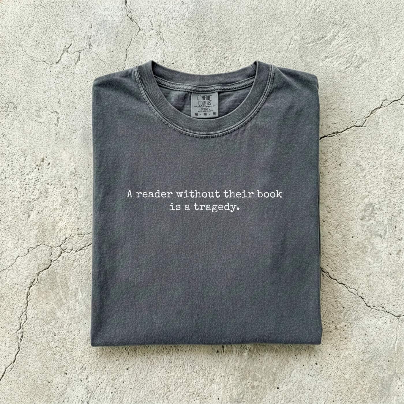 Pepper Comfort Colors tee folded flat on a concrete background reading "A reader without their book is a tragedy." in a minimal typewriter font.