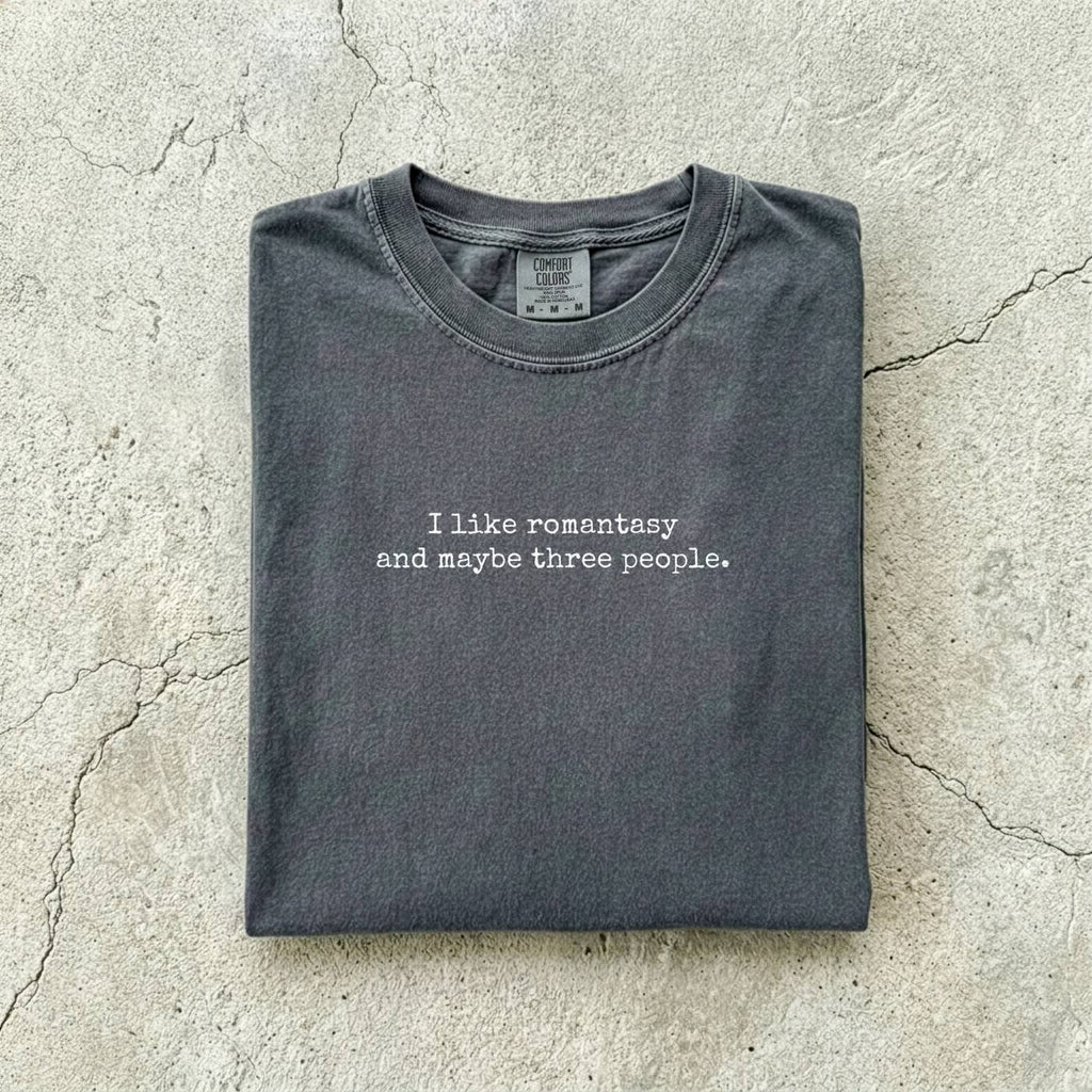 Pepper Comfort Colors tee folded flat reading "I like romantasy and maybe three people." in a typewriter font.