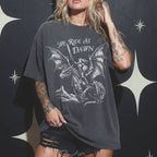 Woman wearing a pepper color relaxed fit Comfort Colors tee with a vintage graphic of a woman warrior dragon rider and "We Ride at Dawn" text.