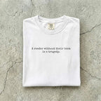 White Comfort Colors tee folded flat on a concrete background reading "A reader without their book is a tragedy." in a minimal typewriter font.