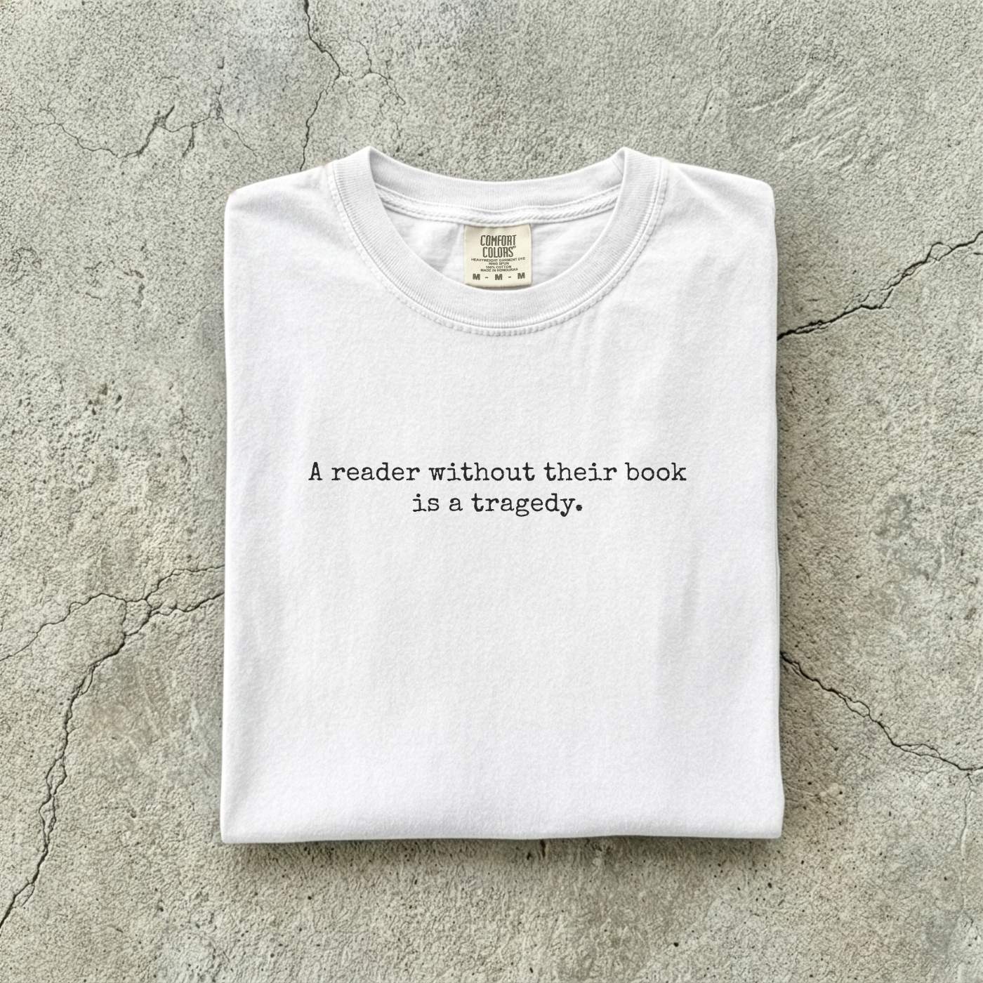 White Comfort Colors tee folded flat on a concrete background reading "A reader without their book is a tragedy." in a minimal typewriter font.