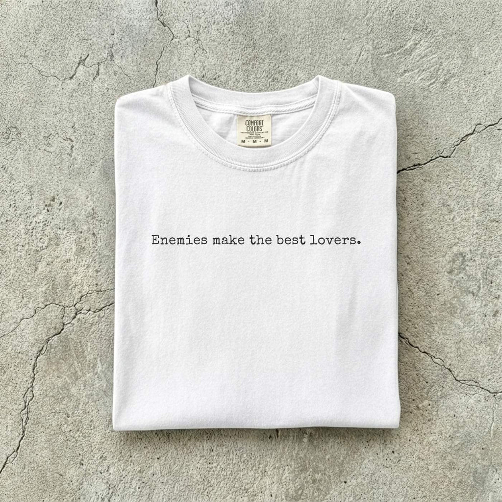 White Comfort Colors tee folded flat on a concrete background reading "Enemies make the best lovers." in a minimal typewriter font.