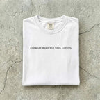 White Comfort Colors tee folded flat on a concrete background reading "Enemies make the best lovers." in a minimal typewriter font.