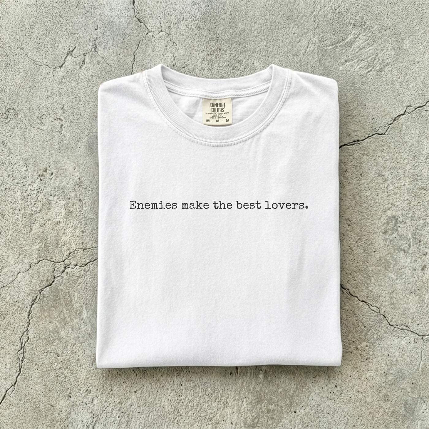 White Comfort Colors tee folded flat on a concrete background reading "Enemies make the best lovers." in a minimal typewriter font.