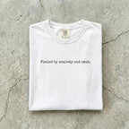 White Comfort Colors tee folded flat on a concrete background reading "Fueled by anxiety and smut." in minimal typewriter font.