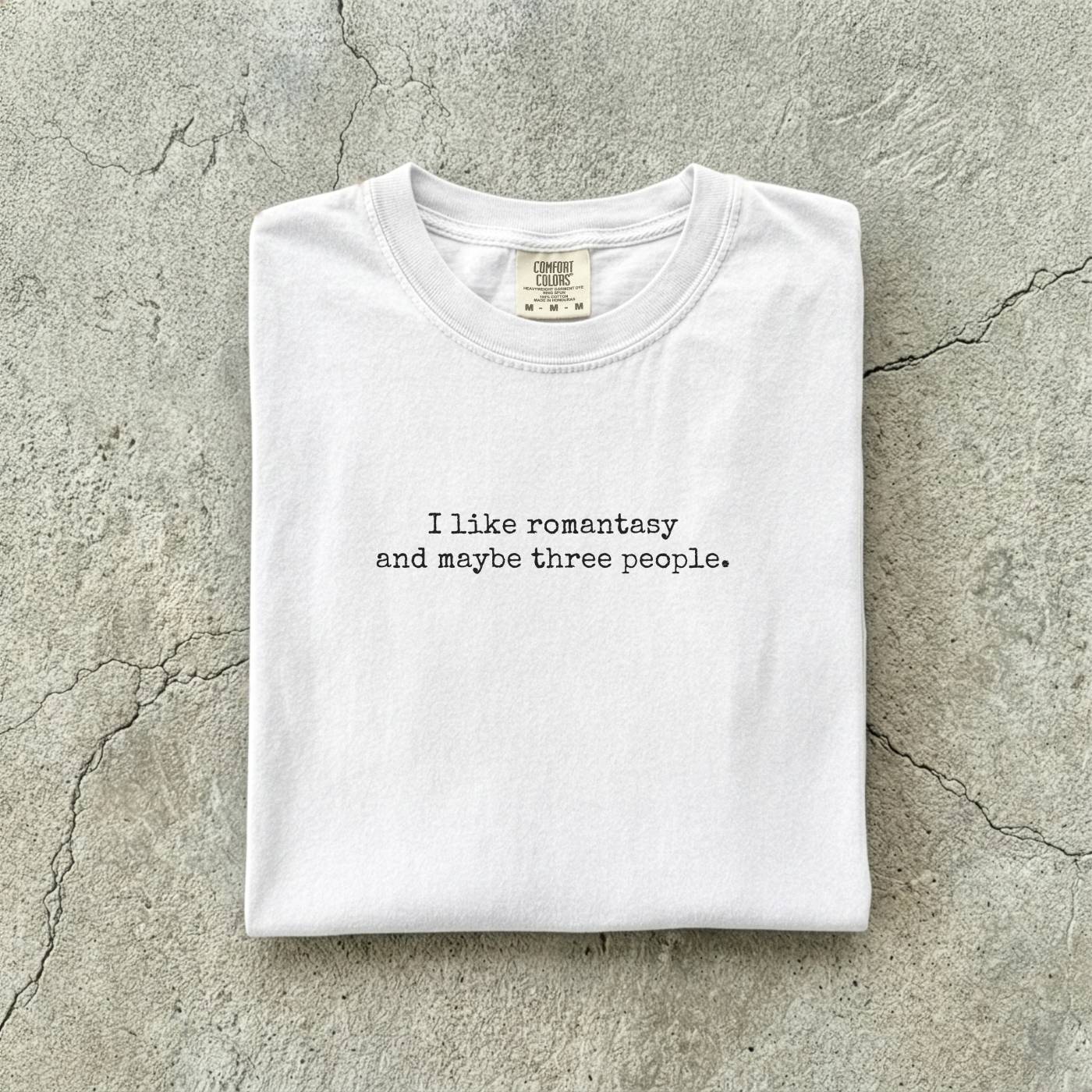 White Comfort Colors tee folded flat reading "I like romantasy and maybe three people." in a typewriter font.