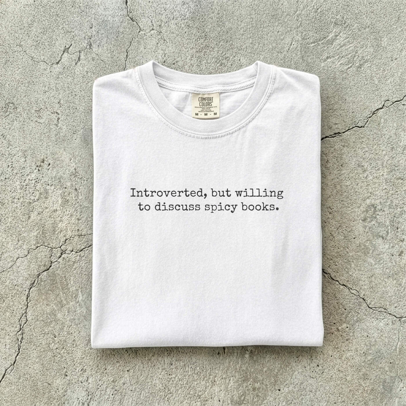 White Comfort Colors tee folded flat on a concrete background reading "Introverted, but willing to discuss spicy books." in a minimal typewriter font.