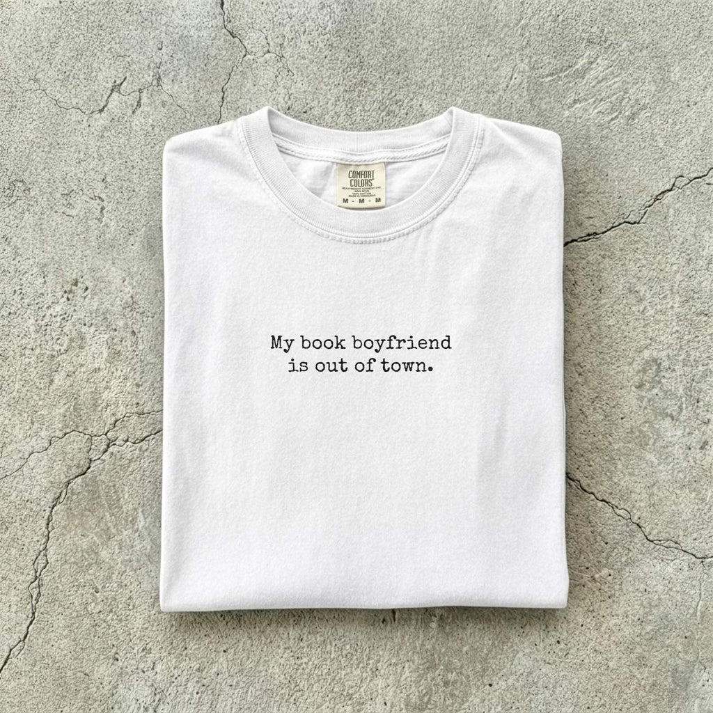 White Comfort Colors tee folded flat on a concrete background reading "My book boyfriend is out of town." in a minimal typewriter font.