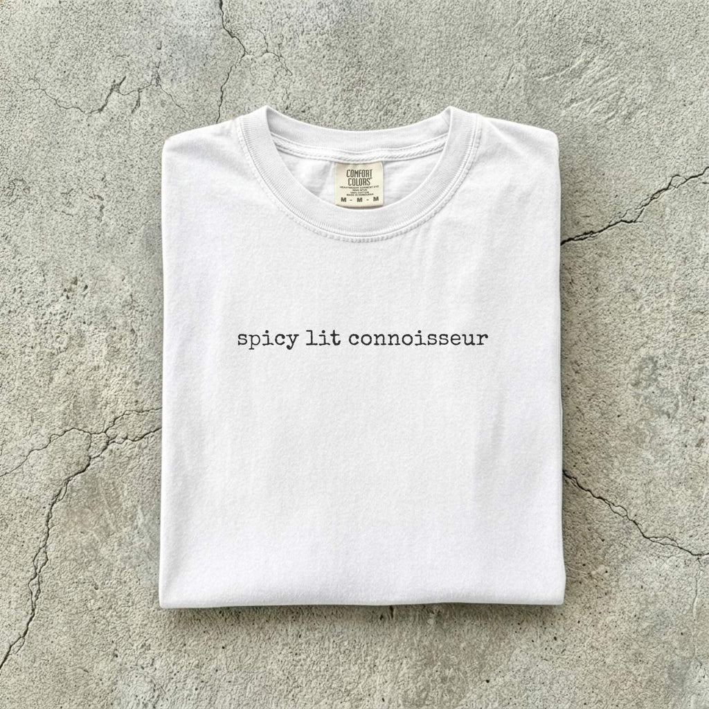 White Comfort Colors tee folded flat on a concrete background reading "spicy lit connoisseur" in a minimal typewriter font.
