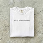White Comfort Colors tee folded flat on a concrete background reading "spicy lit connoisseur" in a minimal typewriter font.