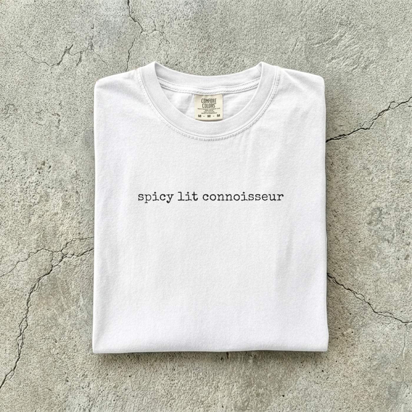 White Comfort Colors tee folded flat on a concrete background reading "spicy lit connoisseur" in a minimal typewriter font.