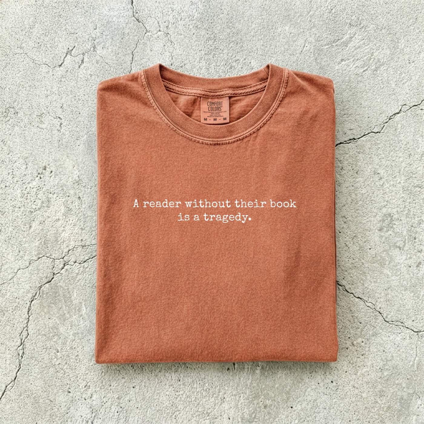 Yam Comfort Colors tee folded flat on a concrete background reading "A reader without their book is a tragedy." in a minimal typewriter font.