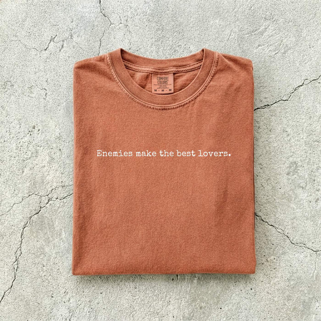 Yam Comfort Colors tee folded flat on a concrete background reading "Enemies make the best lovers." in a minimal typewriter font.