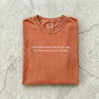 Yam Comfort Colors tee folded flat on a concrete background reading "Introverted, but willing to discuss spicy books." in a minimal typewriter font.