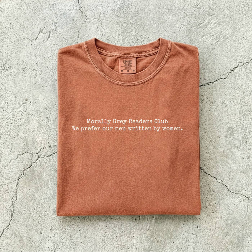 Yam Comfort Colors tee folded flat on a concrete background reading "Morally grey readers club. We prefer our men written by women." in a minimal typewriter font.